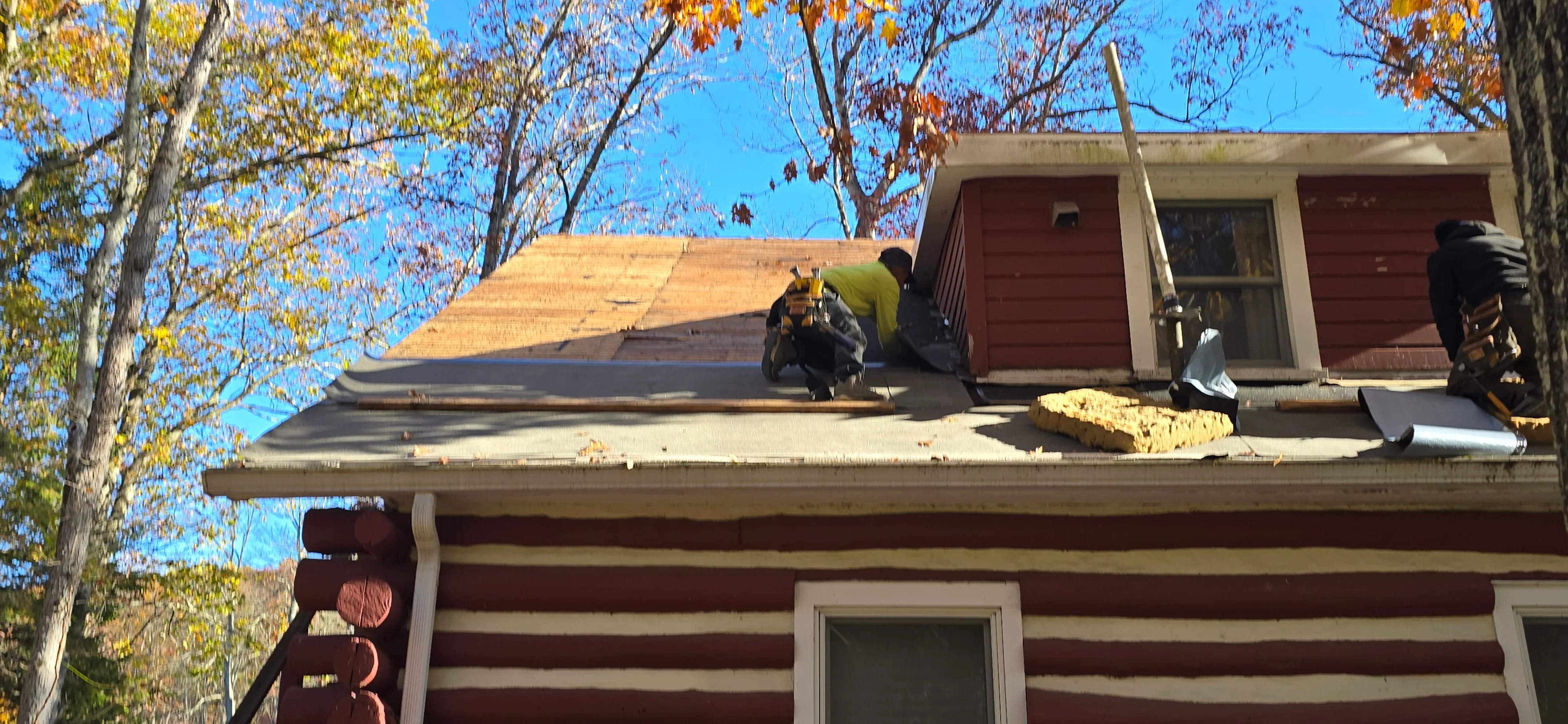 Roofers installing new asphalt shingles on pitched residential roof with autumn trees visible